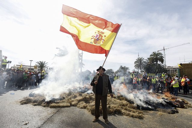 A protestor waves a Spanish flag as hay burns during a farmers protest to denounce their conditions and the European agricultural policy, aiming to block the port of Valencia, on February 22, 2024. Spanish farmers and livestock breeders have been protesting since February 1 for product prices to cover their production costs, and for non-EU imports to face the same regulations they do. Spanish farmers are part of a wider movement that has seen roads blocked in France, Germany, Italy, Poland and Greece, among other countries. (Photo by Jose Jordan/AFP Photo)