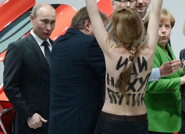 A topless demonstrator with written messages on her back walks towards Russian President Vladimir Putin, left and German Chancellor Angela Merkel, right, during the opening tour at the Hannover Fair in Hannover, Germany, Monday April 8, 2013. Several activists stormed the booth of Volkswagen to demonstrate in presence of the politicians. (Photo by Jochen Lurbke/AP Photo/Dpa)