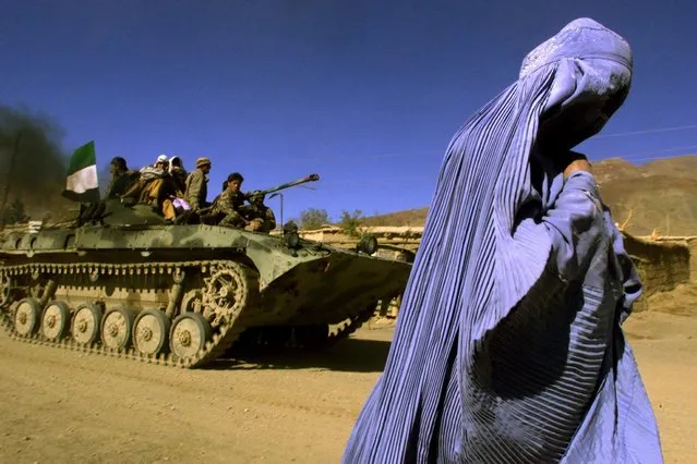 An Afghan woman wearing a traditional Burqa walks on the side of a road as a Northern Alliance APC, (Armoured Personnel Carrier) carrying fighters and the Afghan flag, drives to a new position in the outskirts of Jabal us Seraj, some 60kms north of the Afghan capital Kabul, November 4, 2001. (Photo by Yannis Behrakis/Reuters)