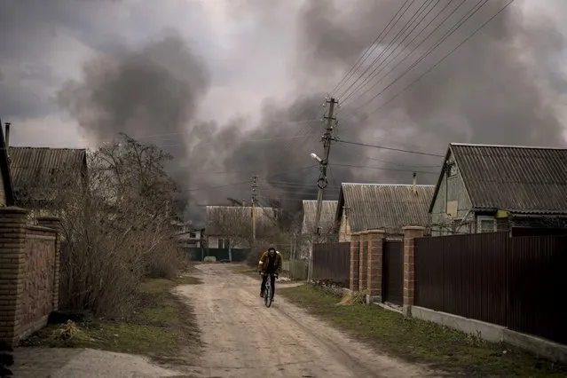 A Ukrainian man rides his bicycle near a factory and a store burning after it had been bombarded in Irpin, on the outskirts of Kyiv, Ukraine, Sunday, March 6, 2022. (Photo by Emilio Morenatti/AP Photo)