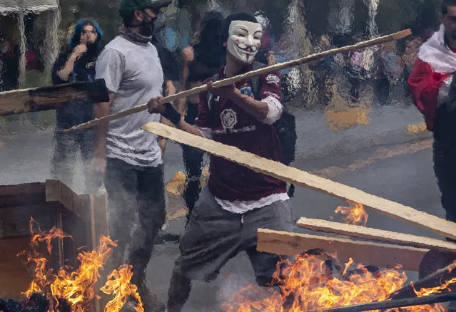 A demonstrator wearing a mask throws a plank of wood into a flaming barricade during a protests in Santiago, Chile, Saturday, October 19, 2019. The protests started on Friday afternoon when high school students flooded subway stations, jumping turnstiles, dodging fares and vandalizing stations as part of protests against a fare hike, but by nightfall had extended throughout Santiago with students setting up barricades and fires at the entrances to subway stations, forcing President Sebastian Pinera to announce a state of emergency and deploy the armed forces into the streets. (Photo by Esteban Felix/AP Photo)