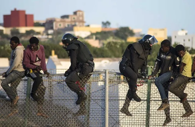 African migrants sit atop a border fence, as Spanish Civil Guard officers in riot gear climb up to force them to climb down during an attempt to cross into Spanish territories, between Morocco and Spain's north African enclave of Melilla October 22, 2014. Around 400 migrants attempted to cross the border into Spain, according to local media. (Photo by Jesus Blasco de Avellaneda/Reuters)