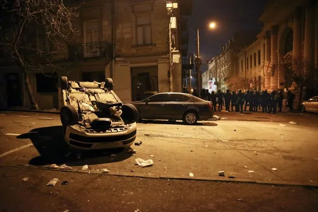 An overturned car is seen in front of a police line that blocked a street to stop protesters outside the Georgian parliament building in Tbilisi, Georgia, early Thursday, March 9, 2023. Police in Georgia's capital have fired water cannons and tear-gas to disperse demonstrators around the parliament building protesting a draft law aimed at curbing the influence of “foreign agents”, among fears it could be used to silence opposition. (Photo by Zurab Tsertsvadze/AP Photo)