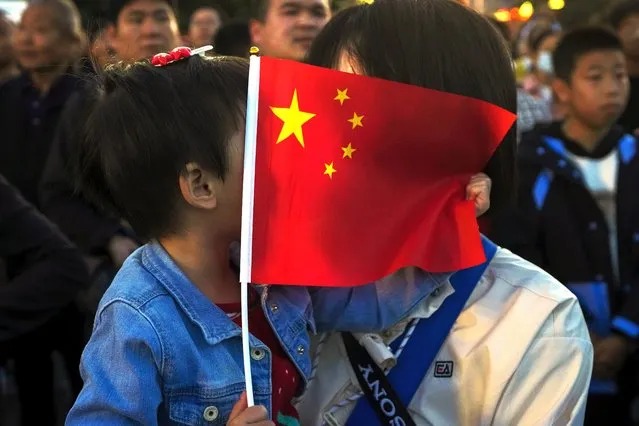 A child talks to her mother behind a national flag as people gather on a street near Tiananmen Square to watch a flag-raising ceremony on the National Day in Beijing, Sunday, Octpber 1, 2023. Hundreds of thousands of people watched a flag-raising ceremony near Tiananmen Square on Sunday morning to celebrate the 74th National Day of the People's Republic of China. (Photo by Andy Wong/AP Photo)