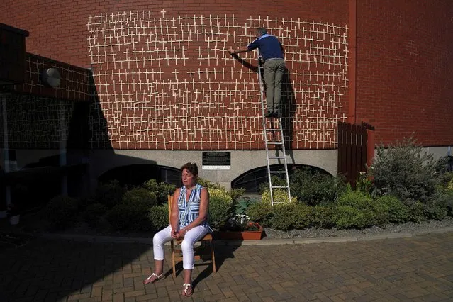 A parishioner prays as Patrick Hand affixes crosses to the outdoor chapel of a thousand crosses and a million tears as at Balally Parish Church of the Ascension of the Lord, following the outbreak of the coronavirus disease (COVID-19), in Dublin, Ireland, May 27, 2020. The outdoor chapel's wall of crosses has become a shrine and each cross represents a person in Ireland that has died from coronavirus and is added to daily. (Photo by Clodagh Kilcoyne/Reuters)