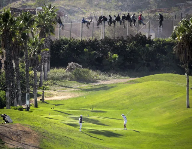 A golfer hits a tee shot as African migrants sit atop a border fence during an attempt to cross into Spanish territories between Morocco and Spain's north African enclave of Melilla October 22, 2014. (Photo by Jose Palazon/Reuters)