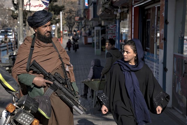 A Taliban fighter stands guard as a woman walks past in Kabul, Afghanistan, Monday, December 26, 2022. Recent Taliban rulings on Afghan women include bans on university education and working for NGOs, sparking protests in major cities. Security in the capital Kabul has intensified in recent days, with more checkpoints, armed vehicles, and Taliban special forces on the streets. Authorities have not given a reason for the tougher security. (Photo by Ebrahim Noroozi/AP Photo)