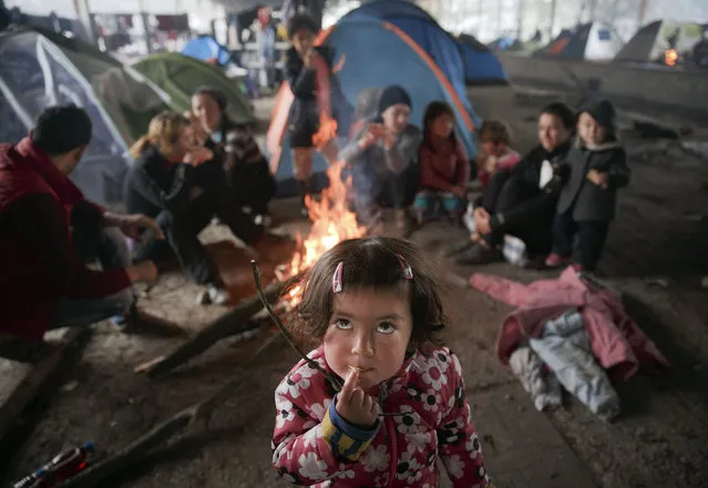 A migrant child eats as others sit around a fire in a railway repairs hangar where people have set up their tents at the northern Greek border point of Idomeni, Greece, Friday, March 18, 2016. (Photo by Vadim Ghirda/AP Photo)