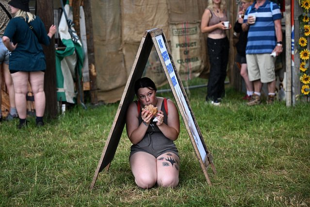 A festivalgoer eats whilst sheltering from the rain beneath an advertising board outside a food stall on the first day of the Glastonbury festival in the village of Pilton, in Somerset, southwest England, on June 21, 2023. The festival takes place from June 21 to June 26. (Photo by Oli Scarff/AFP Photo)