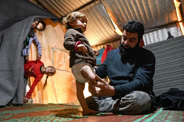 Mohammed, the father of Syrian Maya Merhi, sits next to his disabled toddler, inside the family tent at an Internally Displaced Persons (IDP) camp in Syria's rebel-held northwestern Idlib province, near the Bab al-Hawa border crossing with Turkey, on January 6, 2023. Twelve-year-old Maya and her father were born with no legs due to a congenital condition. She had to struggle around the camp on artificial limbs made of plastic tubing and tin cans. Later, Maya was able to walk thanks to her new prosthetics after undergoing treatment in Turkey. She now has a little brother who is also disabled. (Photo by Rami al Sayed/AFP Photo)