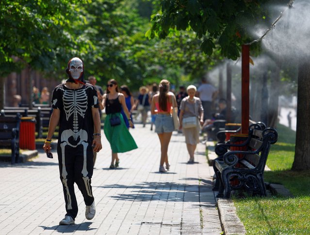 A man handing out promotional leaflets and wearing a skeleton costume walks near a water sprinkler on a hot summer day in central Kyiv, Ukraine on July 10, 2024. (Photo by Valentyn Ogirenko/Reuters)