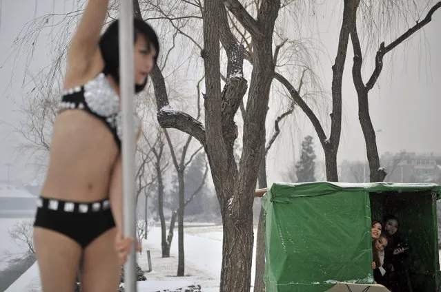 This picture taken on December 17, 2013 shows people (R) sheltering from the cold as they watch a pole dancer (C) practise after it snowed in Tianjin during a promotional event by members of China's national pole dancing team and students of the sport. China set up its first national pole dancing team in Tianjin in 2012 in order to compete in the World Pole Dancing Championships. (Photo by AFP Photo)