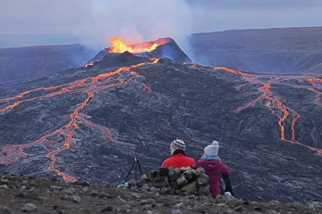 A couple sitting on a hill watch as Fargradalsfjall volcano spews molten lava on August 19, 2021 near Grindavik, Iceland. Iceland is feeling a strong impact from global warming. While the volcano, which erupted in March of this year, lies in the volcanic lowlands southwest of Reykjavik, other Icelandic volcanoes lie under the island's large ice caps, such Eyjafjallajokull, which erupted in 2010. Since the 1990s 90% of Iceland's glaciers have been retreating and projections for the future show a continued and strong retreat in size of its three ice caps. The reduction in mass and pressure from the melting ice caps is increasing the likelihood of further seismic and volcanic activity. (Photo by Sean Gallup/Getty Images)