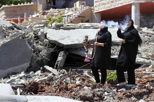 A Lebanese woman smokes a water pipe as another woman flashes a victory sign, while standing on the rubble of a destroyed house that was hit by an Israeli airstrike, in Aita al-Shaab, a Lebanese border village with Israel, south Lebanon, June 29, 2024. (Photo by Mohammed Zaatari/AP Photo)