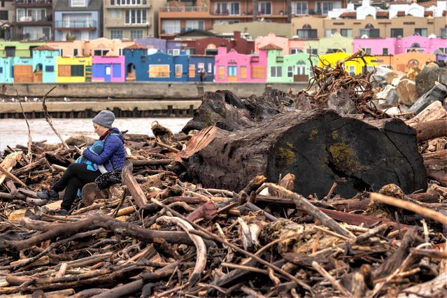 A sits with her baby among trees that were swept into the ocean by recent storms and washed ashore on the beach in Capitola, California, on January 15, 2023. Soggy Californians on Sunday wearily endured their ninth successive storm in a three-week period that has brought destructive flooding, heavy snowfalls and at least 19 deaths, and forecasters said more of the same loomed for another day. (Photo by David McNew/AFP Photo)
