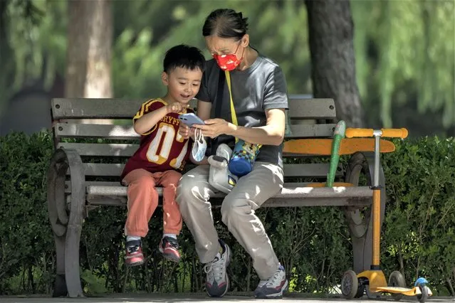 A woman wearing a face mask and a child look at a cellphone as they sit on a bench at a public park in Beijing, on June 2, 2022. As the week-long Lunar New Year holidays in China draw near with promises of feasts and red envelopes stuffed with cash, children have yet another thing to look forward to – one extra hour of online games each day. (Photo by Mark Schiefelbein/AP Photo)