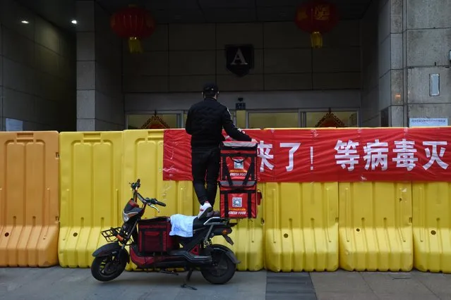 A delivery worker standing on his electric bicycle passes food to customers over barriers built to block an entrance of a residential apartment in Wuhan, Hubei province, China on February 26, 2020. (Photo by Reuters/China Stringer Network)