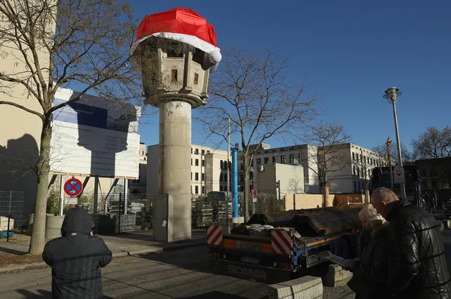 Visitors stand near a former communist-era East German guard tower that has been decorated with an oversized Christmas cap on December 2, 2016 in Berlin, Germany. The museum owner that maintains the tower reportedly intended the cap as a bit of Christmas humour, though not all Berliners think it's funny and charge the move trivializes the deadly history of the Berlin Wall. The guard tower stands on the original spot near Potsdamer Platz where it stood overlooking the Berlin Wall that divded the city between capitalist west and communist east. At least 150 people were killed by East German border guards while trying to flee to the West during the wall's 28-year history. (Photo by Sean Gallup/Getty Images)