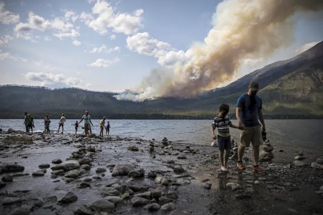 In this Sunday, August 12, 2018 photo provided by the National Park Service, people walk along the shore near Lake McDonald Lodge as the Howe Ridge Fire burns in Glacier National Park, Mont. A wildfire destroyed structures and forced evacuations Monday from the busiest area of Montana's Glacier National Park, as officials in California prepared to reopen Yosemite National Park Tuesday following a nearly three-week closure at the height of the summer season. (Photo by National Park Service via AP Photo)