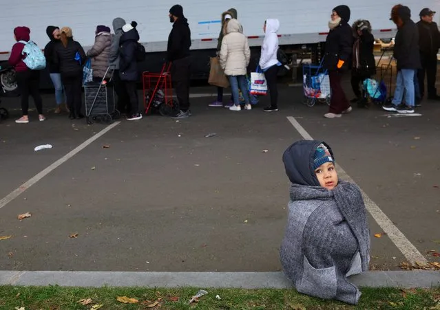 Two year-old Isaac Ospina sits on curb while adults wait in line for free groceries from La Colaborativa's food pantry in Chelsea, Massachusetts on November 8, 2023. (Photo by Brian Snyder/Reuters)