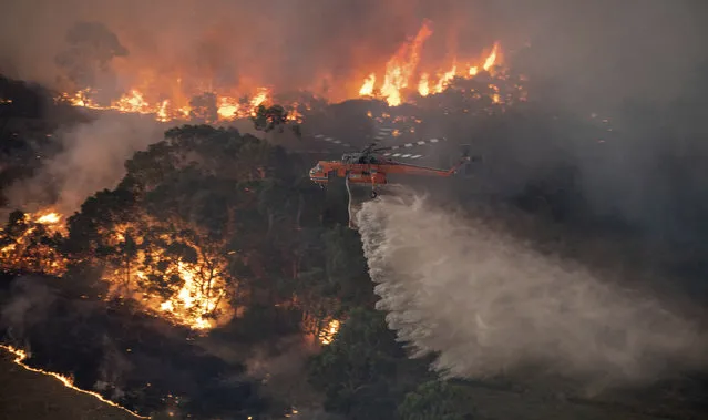 In this Monday, December 30, 2019 photo provided by State Government of Victoria, a helicopter tackles a wildfire in East Gippsland, Victoria state, Australia. Wildfires burning across Australia's two most-populous states trapped residents of a seaside town in apocalyptic conditions Tuesday, Dec. 31, and were feared to have destroyed many properties and caused fatalities. (Photo by State Government of Victoria via AP Photo)