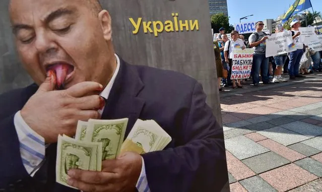 People rally near a placard depicting a banker counting money set in front of Ukrainian parliament in Kiev during protest action of borrowers of Ukrainian banks on July 1, 2014. The borrowers, who took credits in the western currency for the construction or purchase of housing, protest against the policy of the banks which make enormous profits at the expense of borrowers after the depreciation of the Ukrainian currency, they claim. (Photo by Sergei Supinsky/AFP Photo)