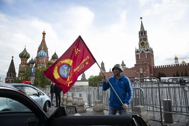 A man poses for a photo with a red flag in front of the closed Red Square during the 75th anniversary of the Nazi defeat in World War II in Moscow, Russia, Saturday, May 9, 2020. Victory Day is Russia's most important secular holiday and this year's observance had been expected to be especially large because it is the 75th anniversary, but the Red Square military parade and a mass procession called The Immortal Regiment were postponed as part of measures to stifle the spread of coronavirus. (Photo by Pavel Golovkin/AP Photo)