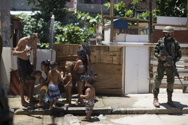 Residents line up to take showers in an alleyway as a soldier patrols during a surprise operation in Jacarezinho slum in Rio de Janeiro, Brazil, Thursday, January 18, 2018. Troops have been sent to Rio due to the increase of the violence, and in an attempt to help restore order, but so far have had little impact. The operation on Thursday took place three weeks ahead of Carnival, when thousands of tourists are expected to arrive in the city. (Photo by Leo Correa/AP Photo)