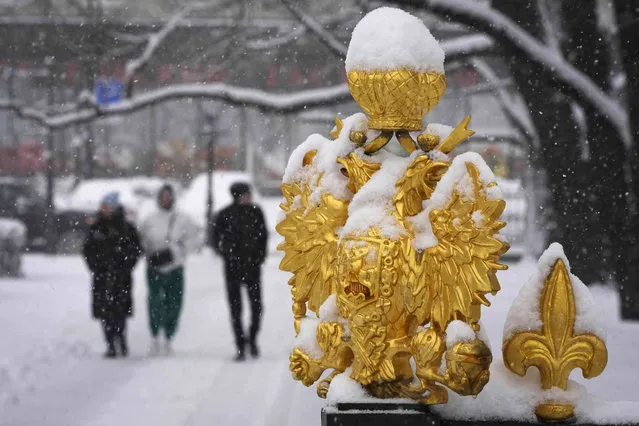People walk in snow covered streets past a double-headed eagle, the coat of arms of Russia, installed on the fence of a park in St. Petersburg, Russia, Wednesday, March 29, 2023. A storm has brought heavy snowfall to St. Petersburg. (Photo by Dmitri Lovetsky/AP Photo)