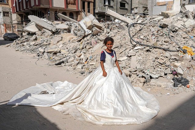 A Palestinian girl poses with a wedding dress found amid the rubble of buildings destroyed during Israeli bombardment, in Khan Yunis in the southern Gaza Strip on June 20, 2024, as the conflict in the Palestinian territory between Israel and Hamas continues. (Photo by Bashar Taleb/AFP Photo)