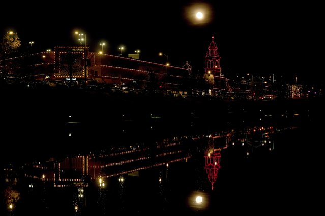 The waning full moon and buildings outlined with Christmas lights is reflected in a creek, Monday, December 16, 2024, in Kansas City, Mo.(Photo by Charlie Riedel/AP Photo)