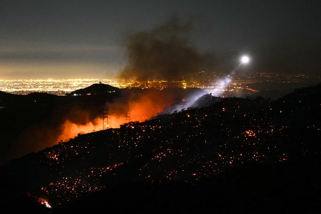 The light of a fire fighting helicopter illuminates a smouldering hillside as the Palisades fire grows near the Mandeville Canyon neighborhood and Encino, California, on January 11, 2025. The Palisades Fire, the largest of the Los Angeles fires, spread toward previously untouched neighborhoods January 11, forcing new evacuations and dimming hopes that the disaster was coming under control. Across the city, at least 11 people have died as multiple fires have ripped through residential areas since January 7, razing thousands of homes in destruction that US President Joe Biden likened to a “war scene”. (Photo by Patrick T. Fallon/AFP Photo)